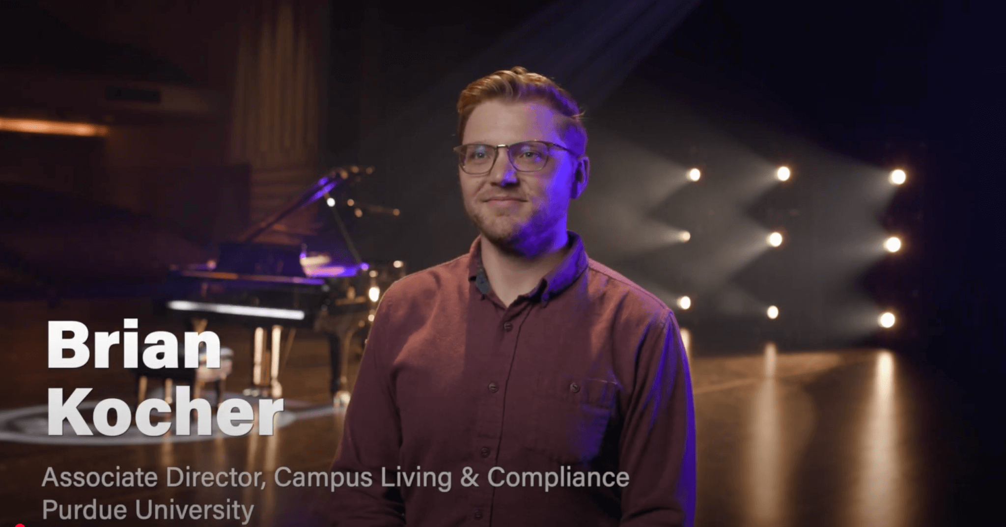 A man with glasses and a trimmed beard stands on a dimly lit stage with spotlights and a grand piano in the background. On-screen text reads, Brian Kocher, Associate Director, Campus Living & Compliance, Purdue University—Purdue Convocations Corporate Sponsors.